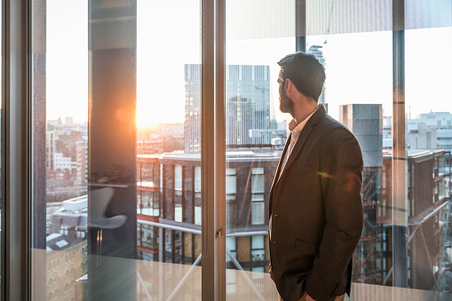 Male office worker looking at view through window of modern office over city. New beginnings, hope, the future. Man looking at sunset.
