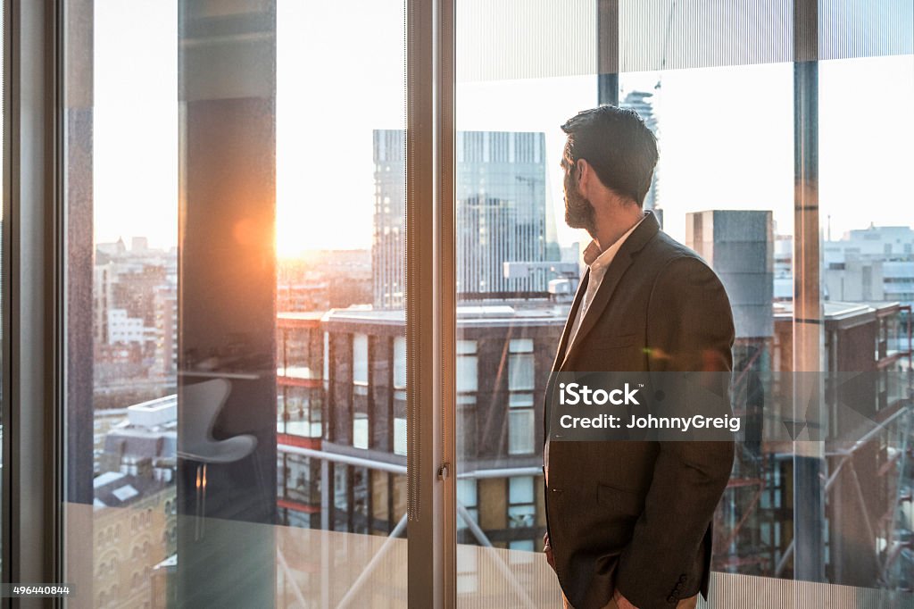 Businessman looking through office window in sunlight Male office worker looking at view through window of modern office over city. New beginnings, hope, the future. Man looking at sunset. Change Stock Photo