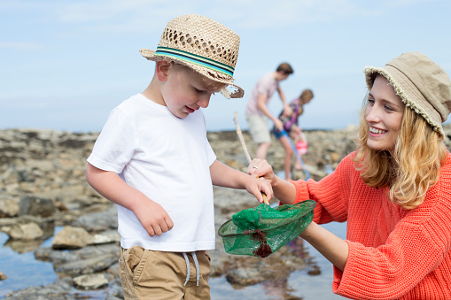 Young family at the beach searching the rock pools. The mother and her young son are in the foreground and she is holding the crab in the net to show her son.