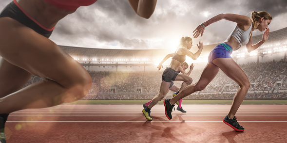 A close up wide angle image of women sprinters running fast during a race in an outdoor athletics track in a generic floodlit arena full of spectators. The athletes are wearing generic running kit and spikes and are competing at an evening track event under a cloudy sky at sunset. 