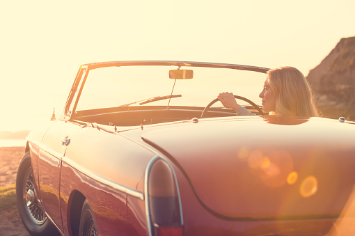 Woman driving a convertible car at the beach. The car is a vintage model shot at sunset or sunrise. The woman looks happy and could be on vacation or having an adventure. Rear view with lens flare. Car can be flipped for Left hand drive counties. 