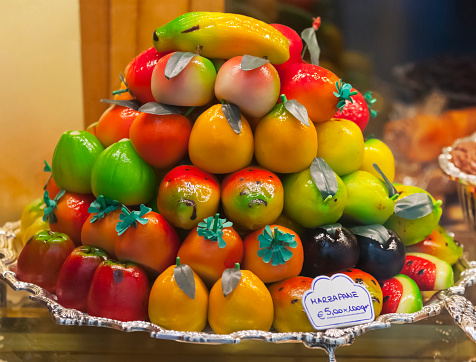 A fruit bowl of marzipan fruits, including, bananas, strawberries, peaches, lemons, oranges, figs, apples, plums, melon, and prickly pears, displayed as a pyramid in a fine patisserie shop window.