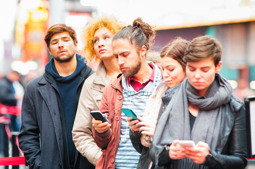 Five young adults waiting in line in Manhattan, some are  using phones to pass the time while the two without phones look exasperated.