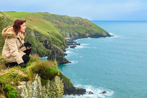 Woman sitting on rock cliff watching the ocean looking to sea. Co. Cork Ireland Europe