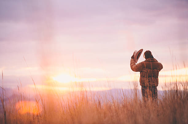 Cowboy holds hat while standing in hay looking at sunset stock photo