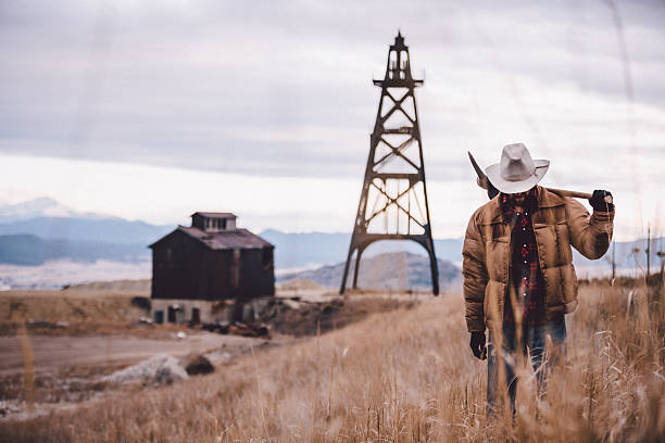 Rancher carries pickaxe through western field mountains in distance