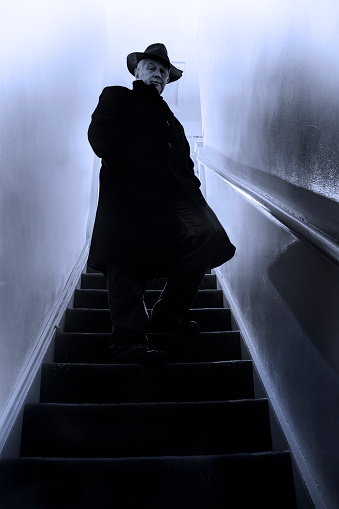 A mysterious man stands midway up the stairs in a stairwell.  The man is cloaked in a long black coat and is wearing a black hat.  The scene is rendered in monochrome, and the man is at the center of the image.  The walls are a light gray color, and there is light flooding into the scene from above.