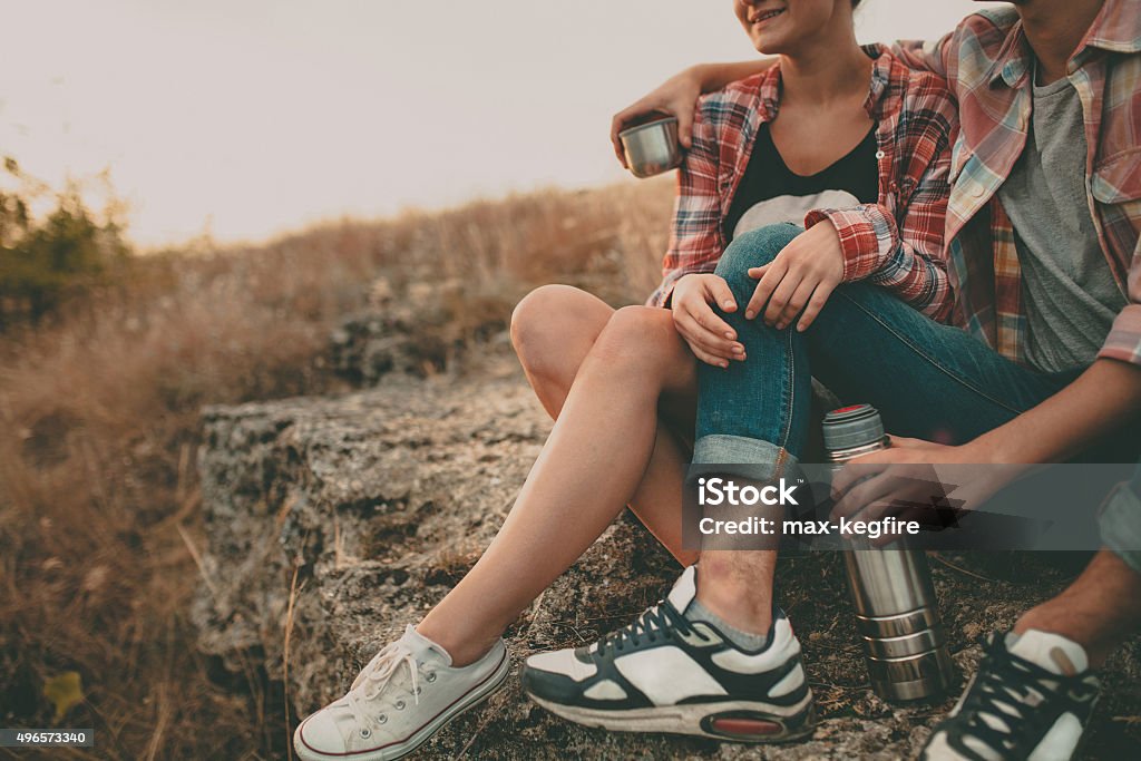 Cropped hipsters having a cup of tea at nature. Cropped shot of young man and woman sitting on a stone with thermos. Teenagers loving couple wearing casual clothes relaxing outdoors 2015 Stock Photo