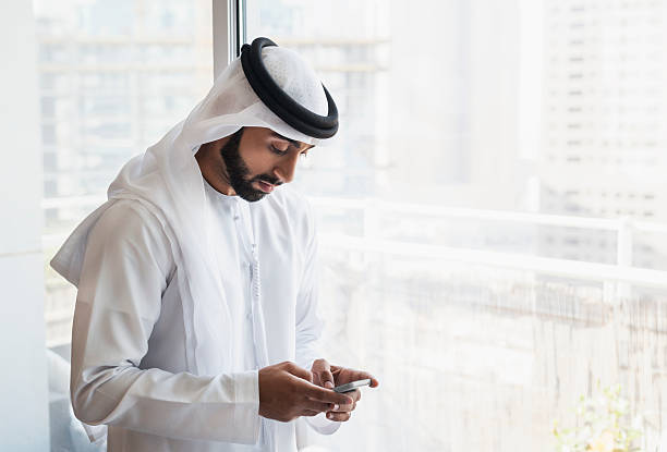 Solitary Arab Man On Phone Inside City Flat Young peaceful Emirati man dressed in traditional clothing kandura, kaffiyeh and agal is texting on his smartphone while standing next to a big transparent window with city skyscrapers background. Image is light, bright, contains copy space on the left and was shot in Dubai, United Arab Emirates. arabian peninsula photos stock pictures, royalty-free photos & images
