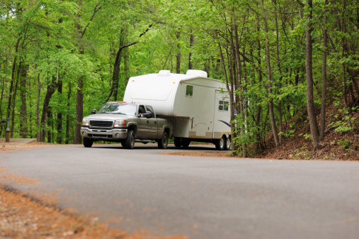 Pickup truck towing fifth wheel recreational vehicle travel trailer up hill on country road in woods.