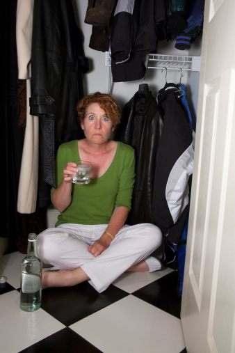 Female hiding in the closet with a bottle and a drink with a surprised expression.