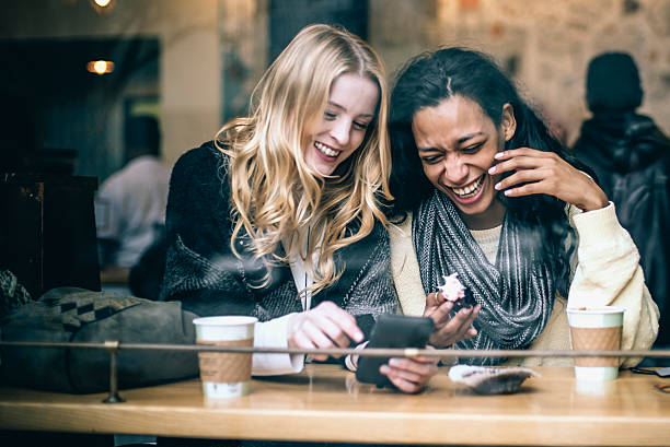 Fun in the coffee shop Two multiethnic young female friends having fun, rejoicing memories using a smart phone while enjoying coffee and pastries in the local NYC coffee shop, natural light, taken from the street. friends laughing stock pictures, royalty-free photos & images