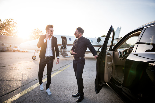 Young man with sunglasses dressed in black standing next to car and opening door to celebity that just arrived by private jet airplane seen in the background. He is also talking over mobile phone.