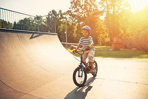 Little boy wearing a helmet is riding a bicycle on ramp in city park on summer day. The boy is aged 6 and is smiling.