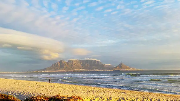 Cape Town's Table Mountain rises from the sea in the late afternoon, draped in her cloudy "Tablecloth"  under a sky filled with dramatic pink and gold tinged clouds. Seen from Blaauwberg beach across Table Bay.