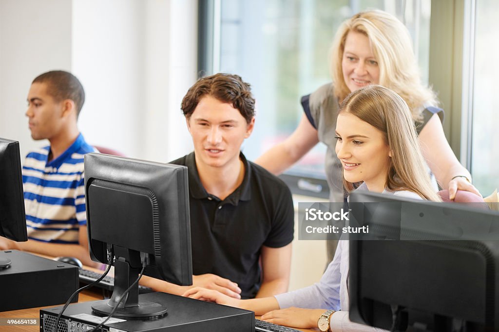 computer class critique female teacher chats to students in the computer lab . She is standing and looking over a student's shoulder   . A male and female student are looking at a shared screen listening to the teacher's advice . 2015 Stock Photo