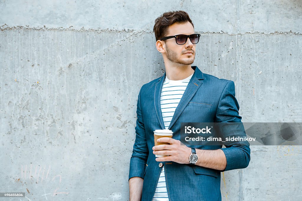 Concept for stylish young man outdoors Portrait of stylish handsome young man with bristle standing outdoors and leaning on wall. Man wearing jacket, sunglasses, shirt and holding cup of coffee Men Stock Photo