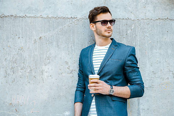 Concept for stylish young man outdoors Portrait of stylish handsome young man with bristle standing outdoors and leaning on wall. Man wearing jacket, sunglasses, shirt and holding cup of coffee well dressed stock pictures, royalty-free photos & images