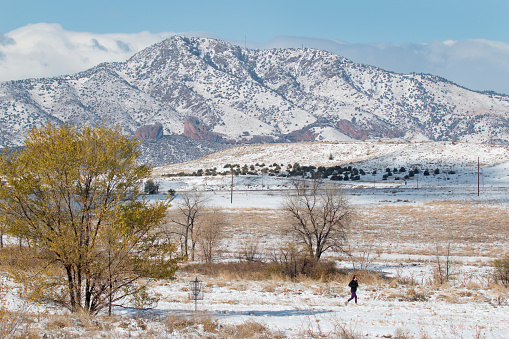 As storm clouds roll past the snow covered Mount Morrison and the Rocky Mountains in the background, a young woman trail runs past a frisbee disc golf course fall colors down a snowy trail in the Fehringer Ranch Park in Littleton, Colorado.
