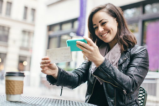 A beautiful Caucasian adult woman sits in a New York city park, taking a picture of a check with her smart phone for a  Remote Deposit Capture.  She smiles, wearing modern stylish clothing with darker and black colors.