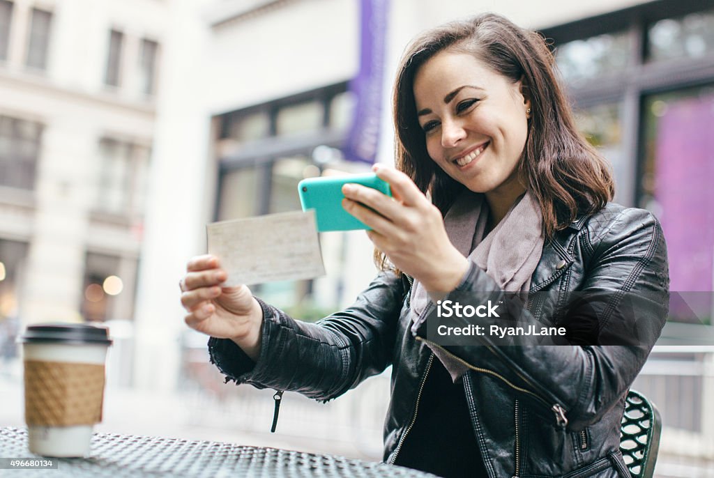New York Woman Depositing Check Remotely A beautiful Caucasian adult woman sits in a New York city park, taking a picture of a check with her smart phone for a  Remote Deposit Capture.  She smiles, wearing modern stylish clothing with darker and black colors. Portable Information Device Stock Photo