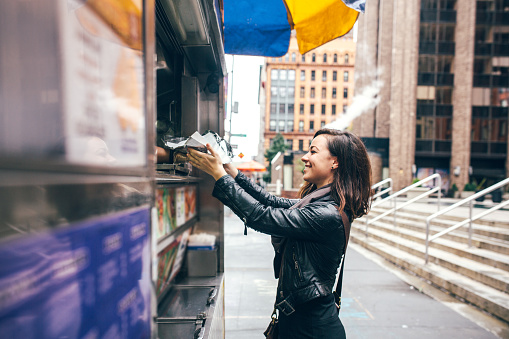 A beautiful Caucasian adult woman buys some lunch from a New York city food cart, a smile on her face.  She wears modern stylish clothing with darker and black colors.  Horizontal image with copy  space.
