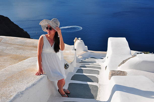 Young beautiful stylish female model tourist lady sitting on the retaining wall enjoying the view in a white dress and blue hat looking at the view of the famous blue domes church and city view the white village of Oia with Caldera and Mediterranean sea panorama travel Greece vacation luxury Europe cruise destination of Thera, Greek Santorini island in Europe.