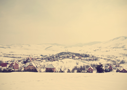 Beautiful idyllic snowy winter landscape in a small picturesque mountain village, on a foggy morning. Image filtered in faded, retro, Instagram style; nostalgic, vintage rural winter concept.