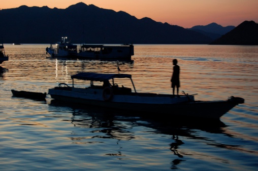 Komodo, Indonesia - November 5, 2013: a young local sailor standing on a wooden boat. In the background the rocky landscape of Komodo island at sunset.