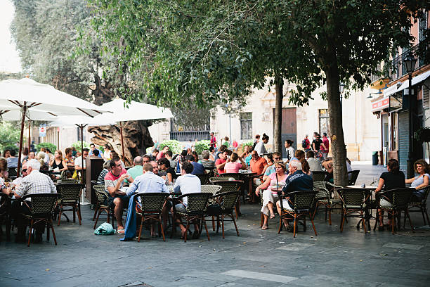 Crowd seated outside restaurant in Palma, Majorca This long shot shows many diverse people sitting in an outdoor dinning area near a restaurant in Palma, Majorca.  The tables are covered by tall, white, fabric, sun-shade umbrellas on the left and mature, dark green trees on the right.  The outdoor restaurant has charcoal gray pavement in front and old buildings in the background. huddle stock pictures, royalty-free photos & images