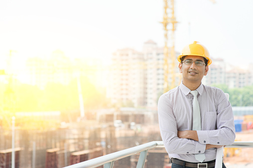 Portrait of Asian Indian male site contractor engineer with hard hat holding blue print paper smiling at construction site, crane with golden sunlight at the background.
