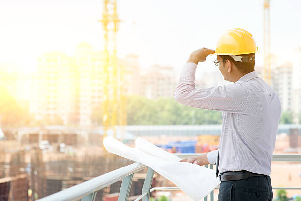 Asian Indian male contractor engineer on site Asian Indian male site contractor engineer with hard hat holding blue print paper looking away inspecting at construction site, crane with golden sunlight at the background. business architecture blue people stock pictures, royalty-free photos & images
