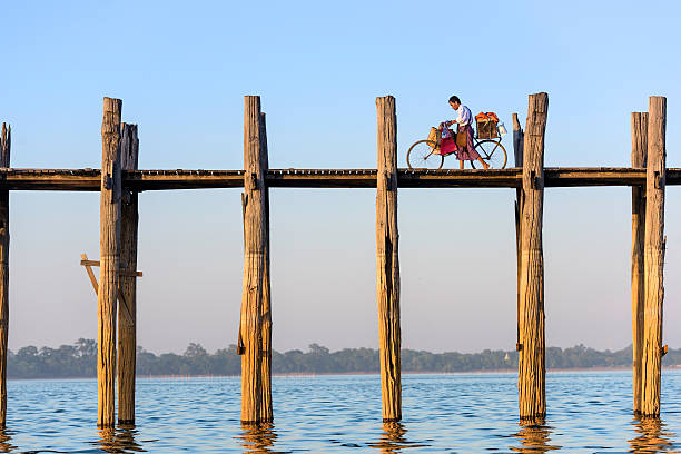 U Bein Bridge Mandalay, Myanmar - October 26, 2015: A cyclist pushes his bike across the U Bein Bridge over Taungthaman Lake. The bridge was built around 1850 and is the oldest and longest teakwood bridge in the world. u bein bridge stock pictures, royalty-free photos & images
