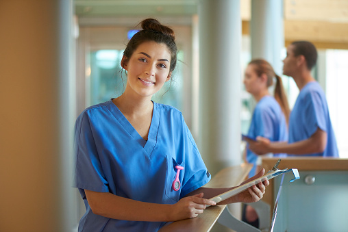 A young female nurse wearing blue scrubs and holding medical records is smiling to camera. She is standing on a busy corridor of a modern hospital . More male and female nurses are walking behind her .