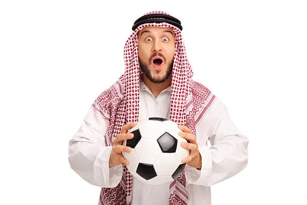 Surprised young Arab in a white robe holding a football and looking at the camera isolated on white background