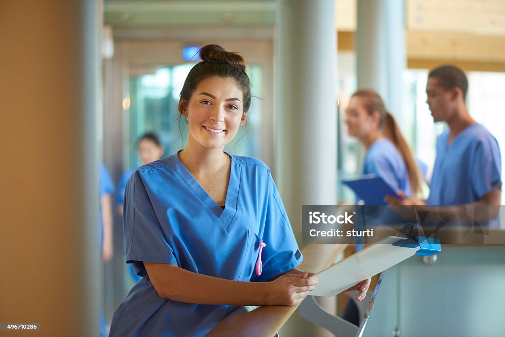 young nurse portrait in hospital A young female nurse wearing blue scrubs and holding medical records is smiling to camera. She is standing on a busy corridor of a modern hospital . More male and female nurses are walking behind her . Nurse Stock Photo young nurse portrait in hospital A young female nurse wearing blue scrubs and holding medical records is smiling to camera. She is standing on a busy corridor of a modern hospital . More male and female nurses are walking behind her . Nurse Stock Photo