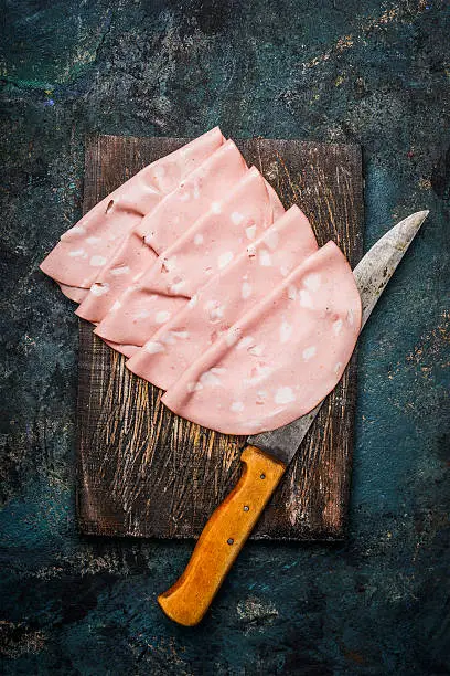 Slices of  Sausage Mortadella di Bologna with kitchen knife on rustic wooden background, top view.