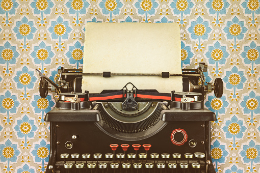 Retro styled image of an old typewriter with a blank sheet of paper in front of wallpaper with a flower print