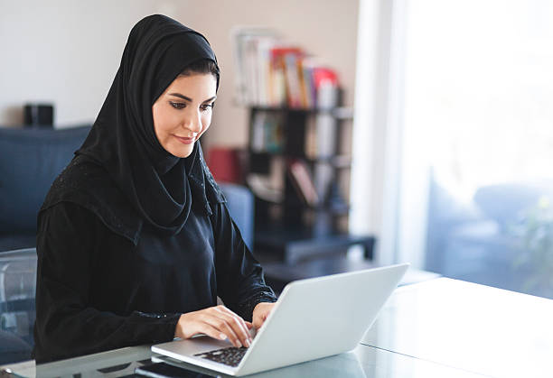 Emirati Woman Working with Laptop at Home Young arabian woman dressed in black religious veil is working on her laptop, looking at screen and typing on keyboard. She seems to be concentrated and glad using various softwares and online services. Image is brightly lit, contains copy space. Made in Dubai, United Arab Emirates. emirati culture photos stock pictures, royalty-free photos & images