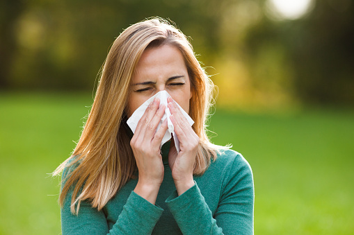 Young woman with allergy symptom blowing nose in park