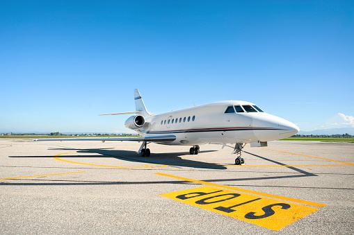 Profile view of stationary private airplane and yellow STOP road sign on asphalt runway against blue clear sky. No people image of air luxury business vehicle, space for copy.
