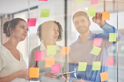 three business colleagues are throwing ideas onto adhesive notes and putting them on the glass wall of their office meeting room .