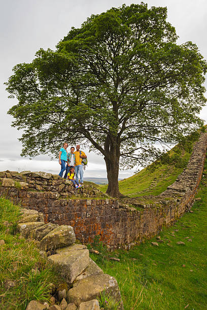An image of a family with two children at Sycamore gap in the countryside. The ancient tree is surrounded by Hadrian's wall in Northumberland, England.