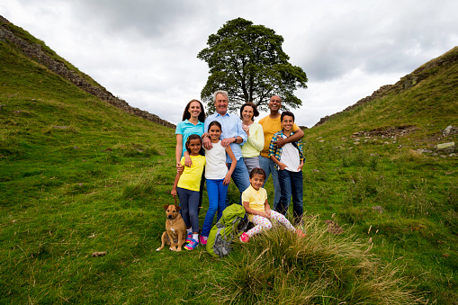 An image of a three generation family posing for a family photograph with their dog past Sycamore gap in the countryside. The ancient tree is surrounded by Hadrian's wall in Northumberland, England. This is a concept shot showing the tree and the three generation family below it.