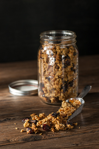 High resolution, digital capture of a metal scoop filled with homemade granola, in front of a jar containing the same. Granola consists of oats, cashews, almonds, dried apricots, dried papaya, dried cranberries, honey, coconut oil, and cinnamon. Scoop and jar sit on a wooden surface in raking light.