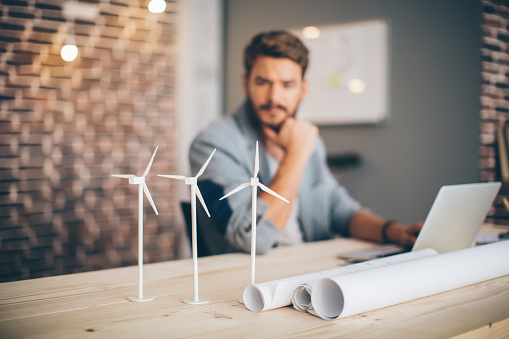 Young engineer at modern office space, sitting at the table and working on energy project.  Focus on wind turbine models on the table.