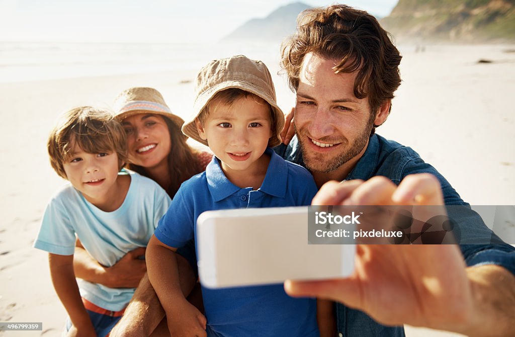 Family taking photo of themselves on beach A young, happy family of four is pictured at the beach, on holiday, taking a selfie shot.  The father is holding a Smartphone out in front of them, as he puts his arm around one of his sons, wearing a barrel hat.  The mother is in the back with her arms around the other son. Beach Stock Photo