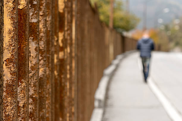 A man goes by the sidewalk next to a rusty fence.