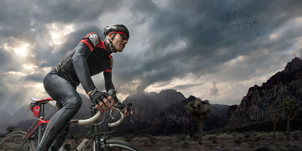 Professional male cyclist cycling on road through mountains at sunset under stormy sky