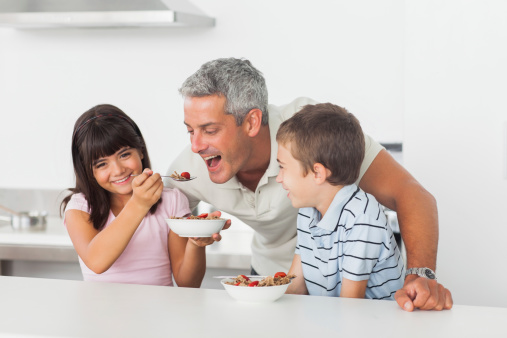 Little girl giving cereal to her father with brother smiling in kitchen